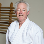 Headshot of Roy May, a white man in his 60s with short gray hair, smiling and wearing a white gi jacket.